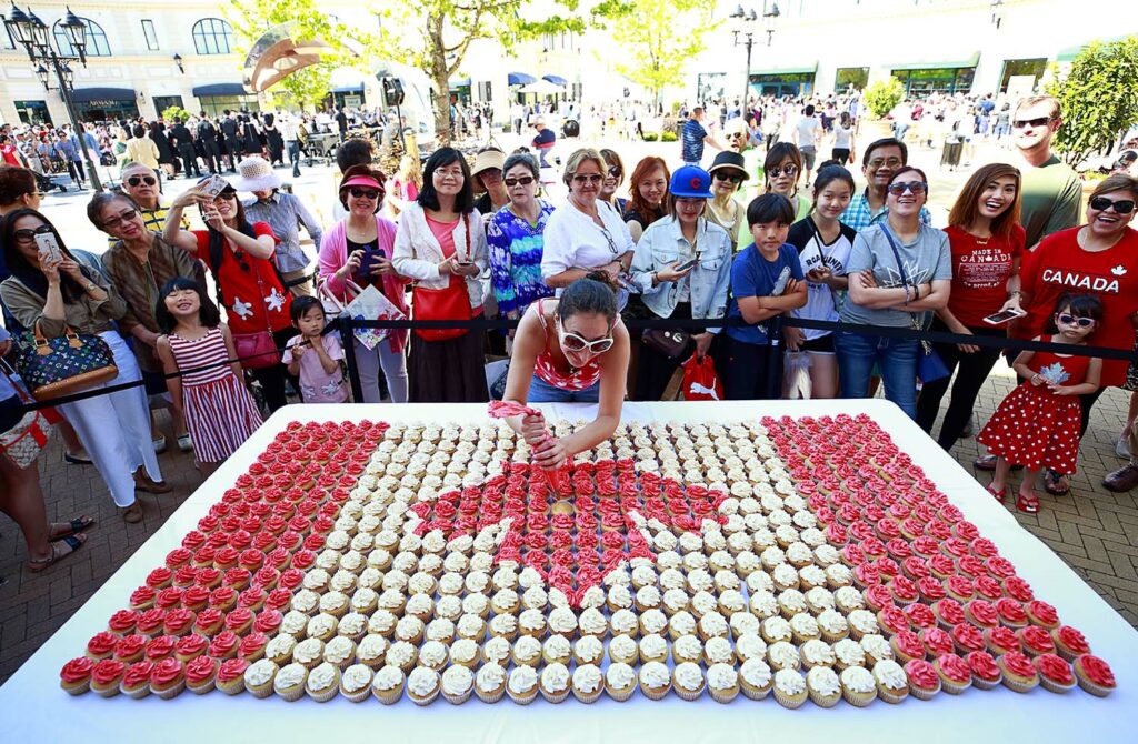 A crowd gathers on a street as a woman decorates many cupcakes with red or white frosting that are arranged to resemble the Canadian flag.