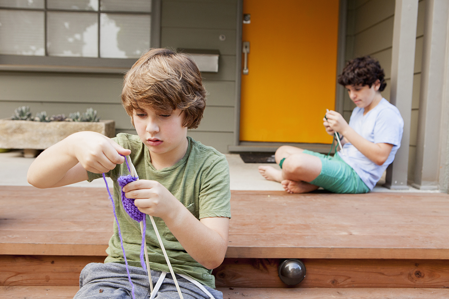 Two boys sit outdoors on a porch and knit.