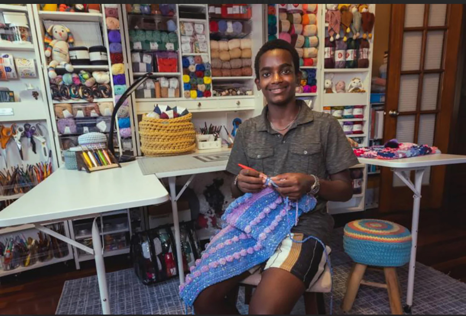 Jonah Larson crochets as he sits in front of shelves of yarn and other crafting supplies.