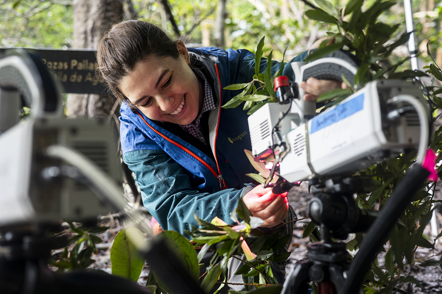 A scientist smiles as she adjusts a machine that is positioned among trees in an outdoor setting.