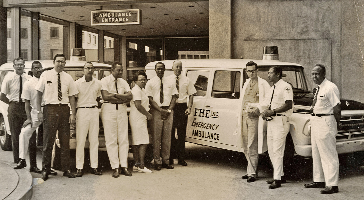 A group of men and women stand in front of an ambulance outside the ambulance entrance of a hospital.
