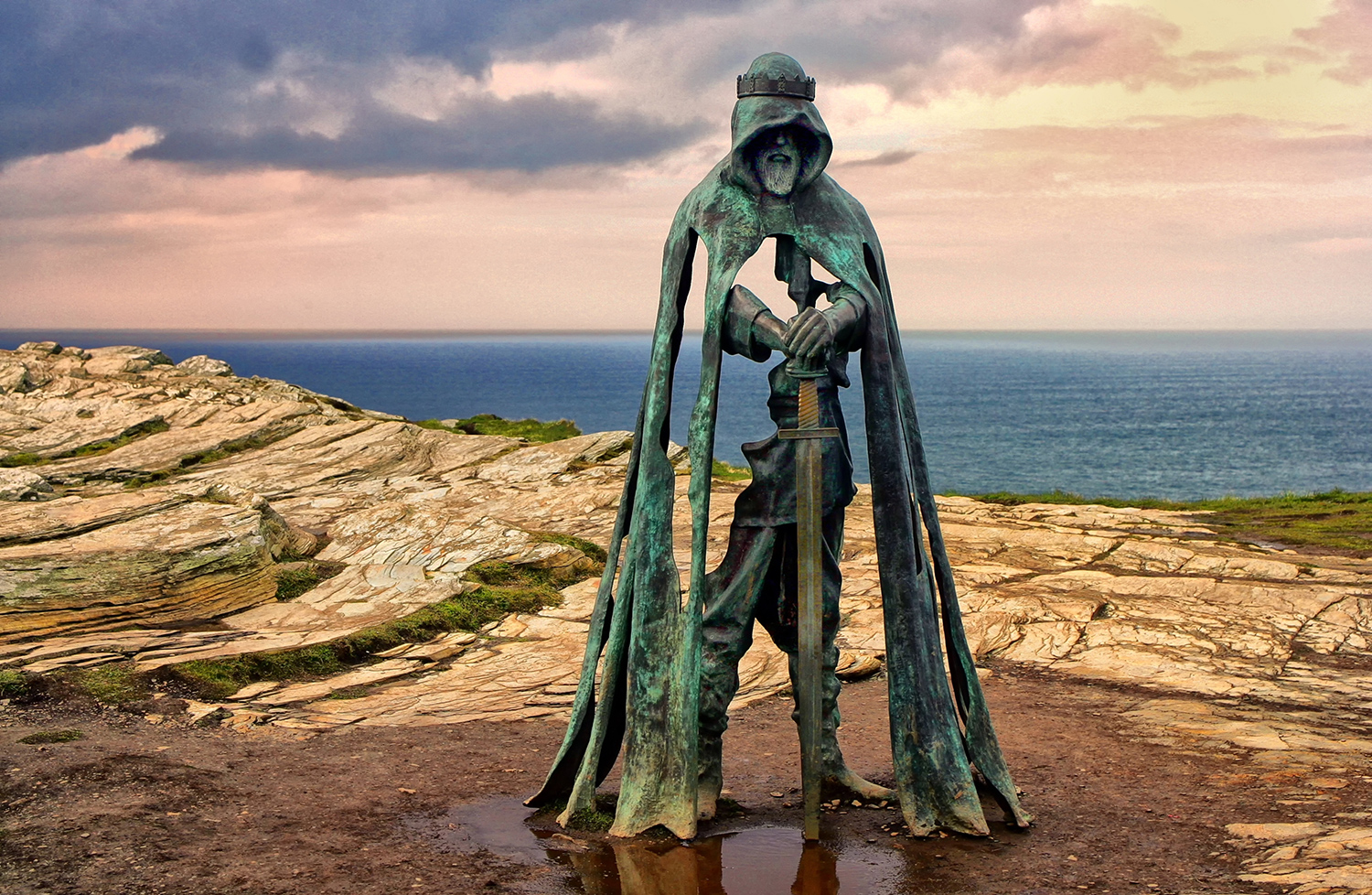 A statue of King Arthur holding a sword is on a cliff overlooking water.
