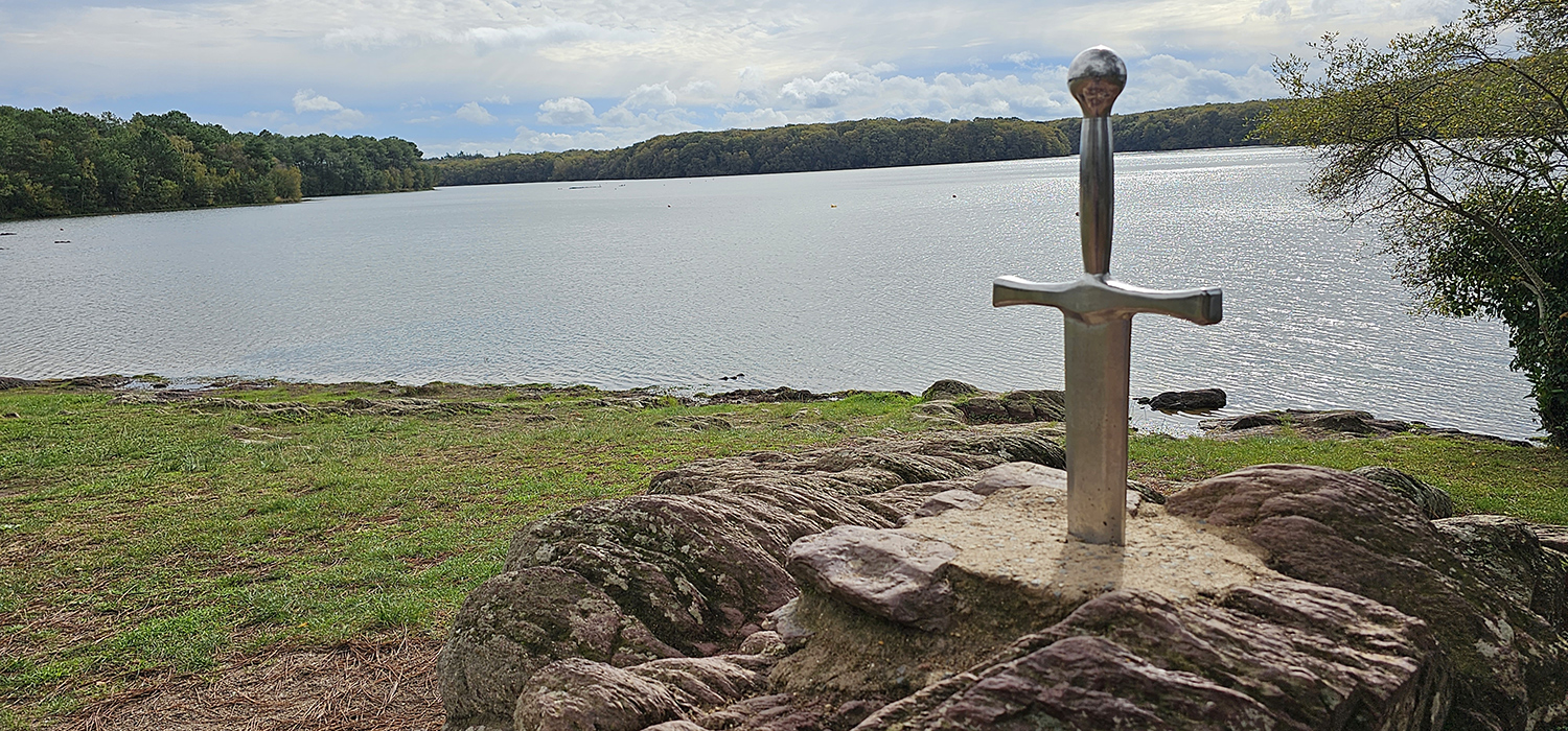 A statue of a sword in a stone with a body of water in the background.