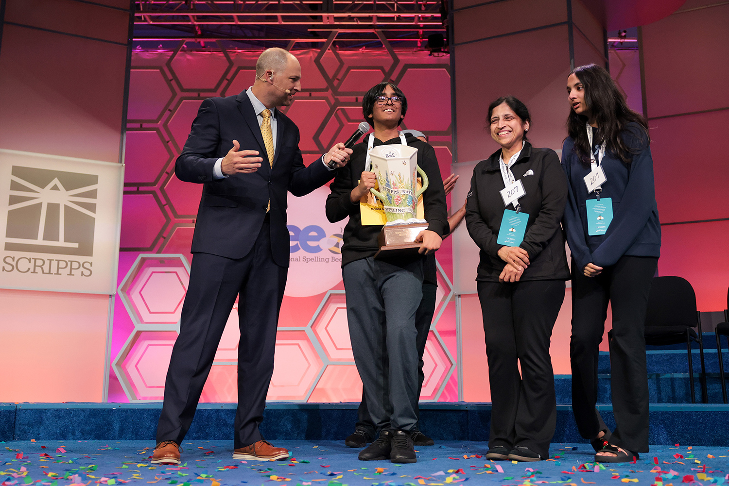 A teenage boy holds a trophy and speaks into a microphone being held by a man as he stands with a woman and a teen girl.