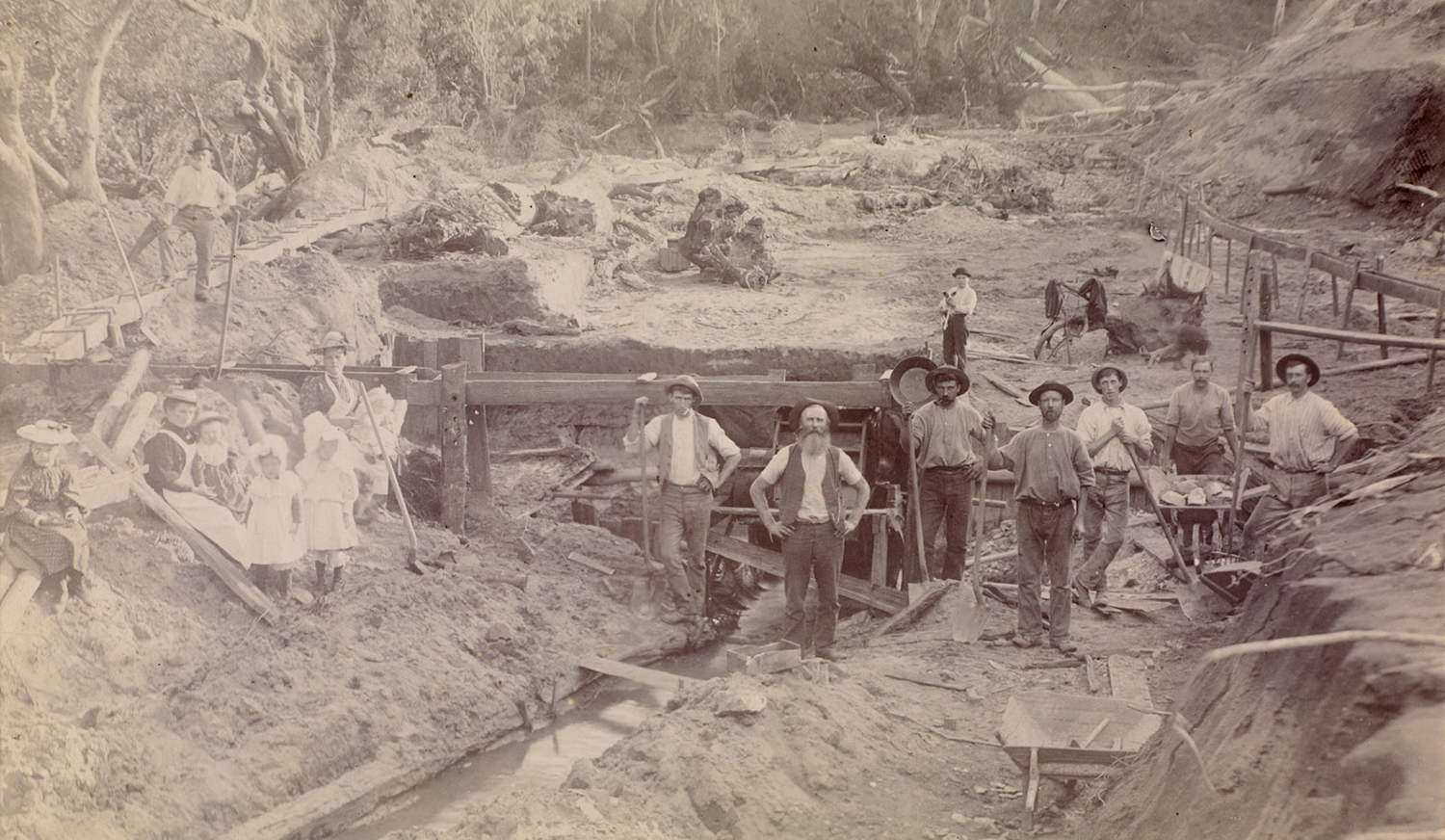 Several men stand at a gold panning site holding tools as two women and some children in 19th century clothing sit on the side.