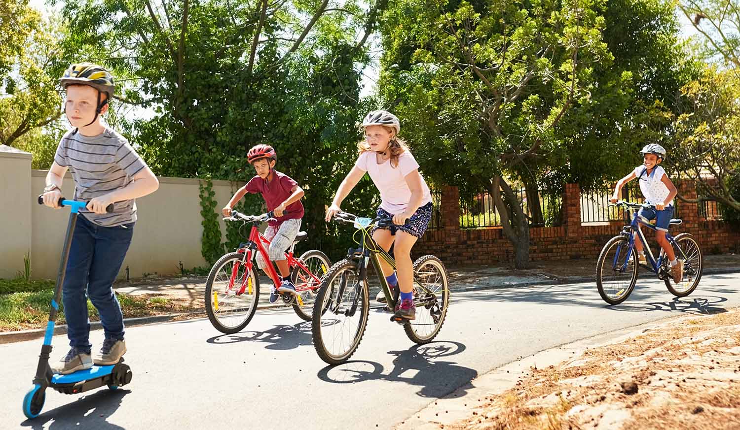 A child on a scooter and three children on bicycles ride down a paved path with trees in the background.