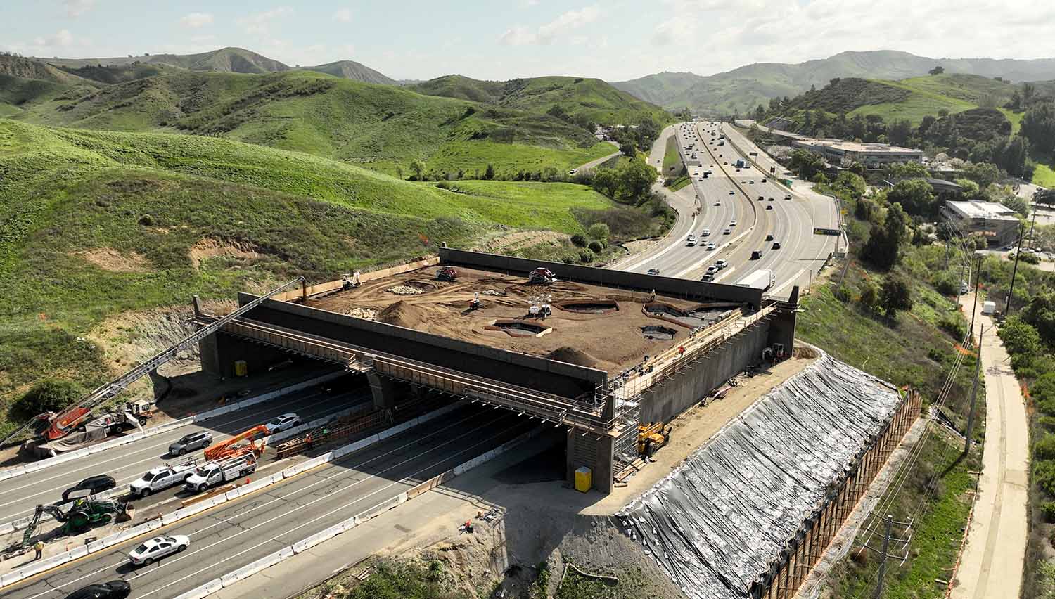 The Wallis Annenberg Crossing under construction over a busy road with green hills in the background.