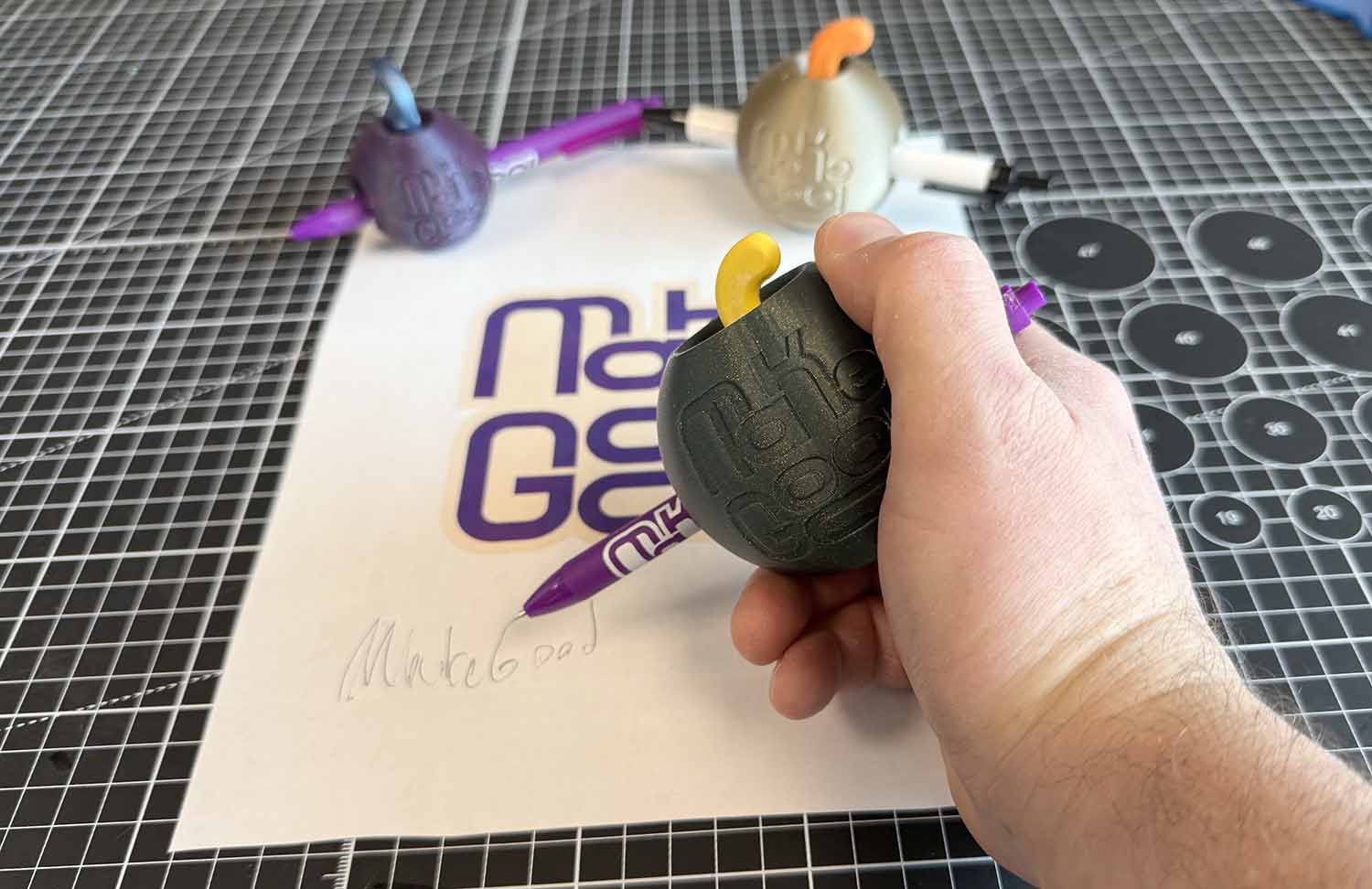 A hand grips a ball that is fitted around a pen and is held over paper where MakeGood has been written.