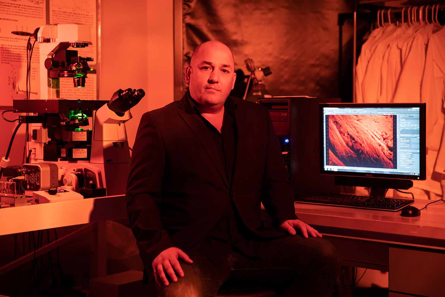 A man poses in a red-lit laboratory where a microscope, lab coats, and other instruments can be seen.