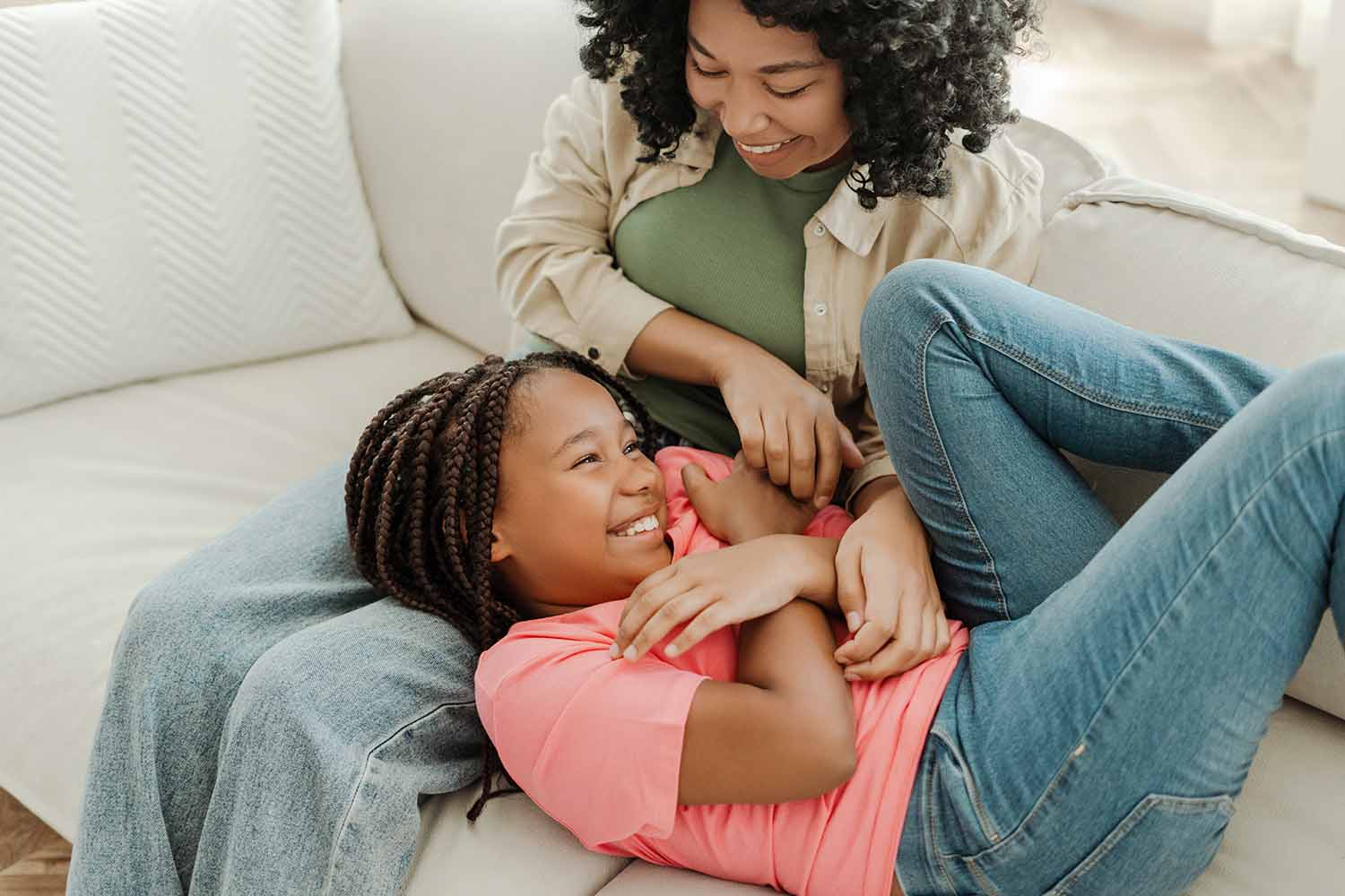 A woman tickles her daughter while sitting on a couch.
