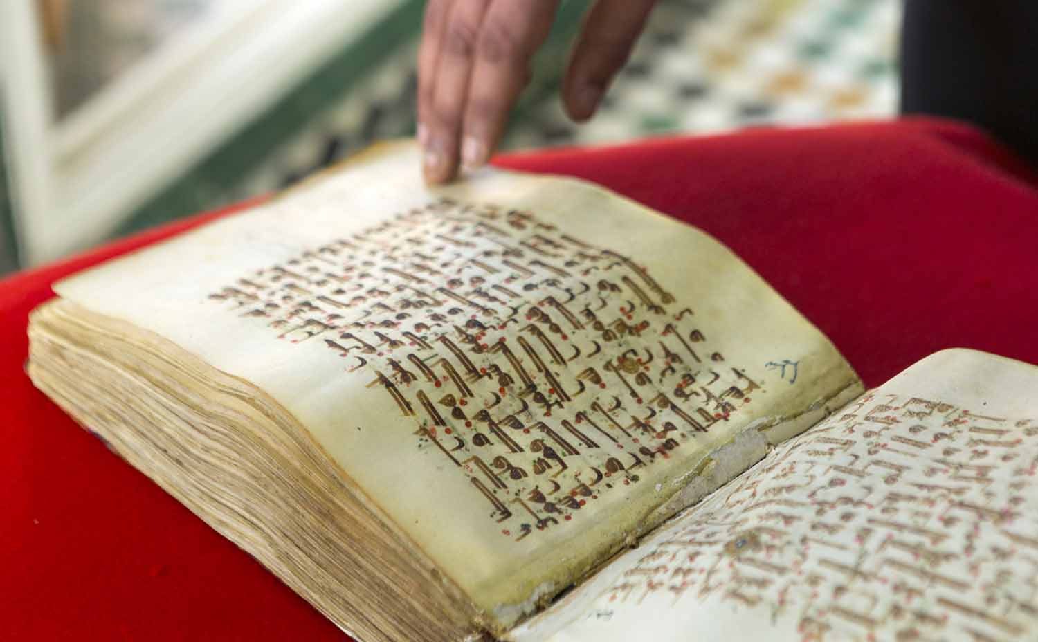An open, handwritten copy of the Koran with fingertips resting on one of the pages.