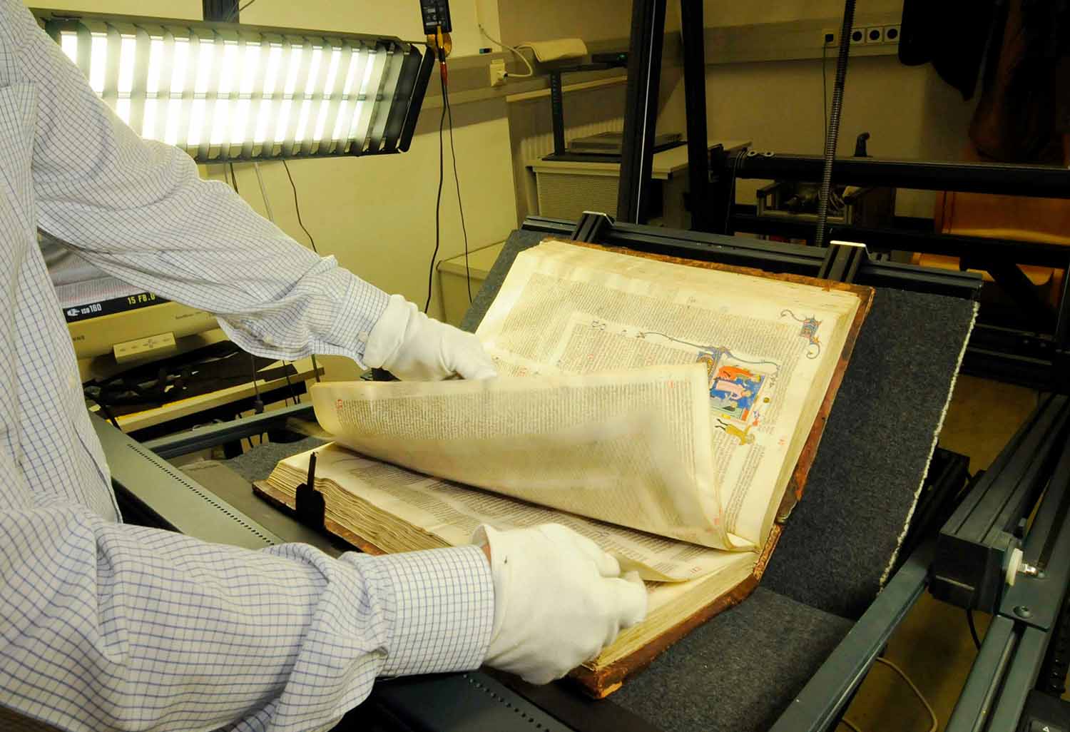 A pair of gloved hands flip through the pages of a very old book that is on a machine, about to be scanned.