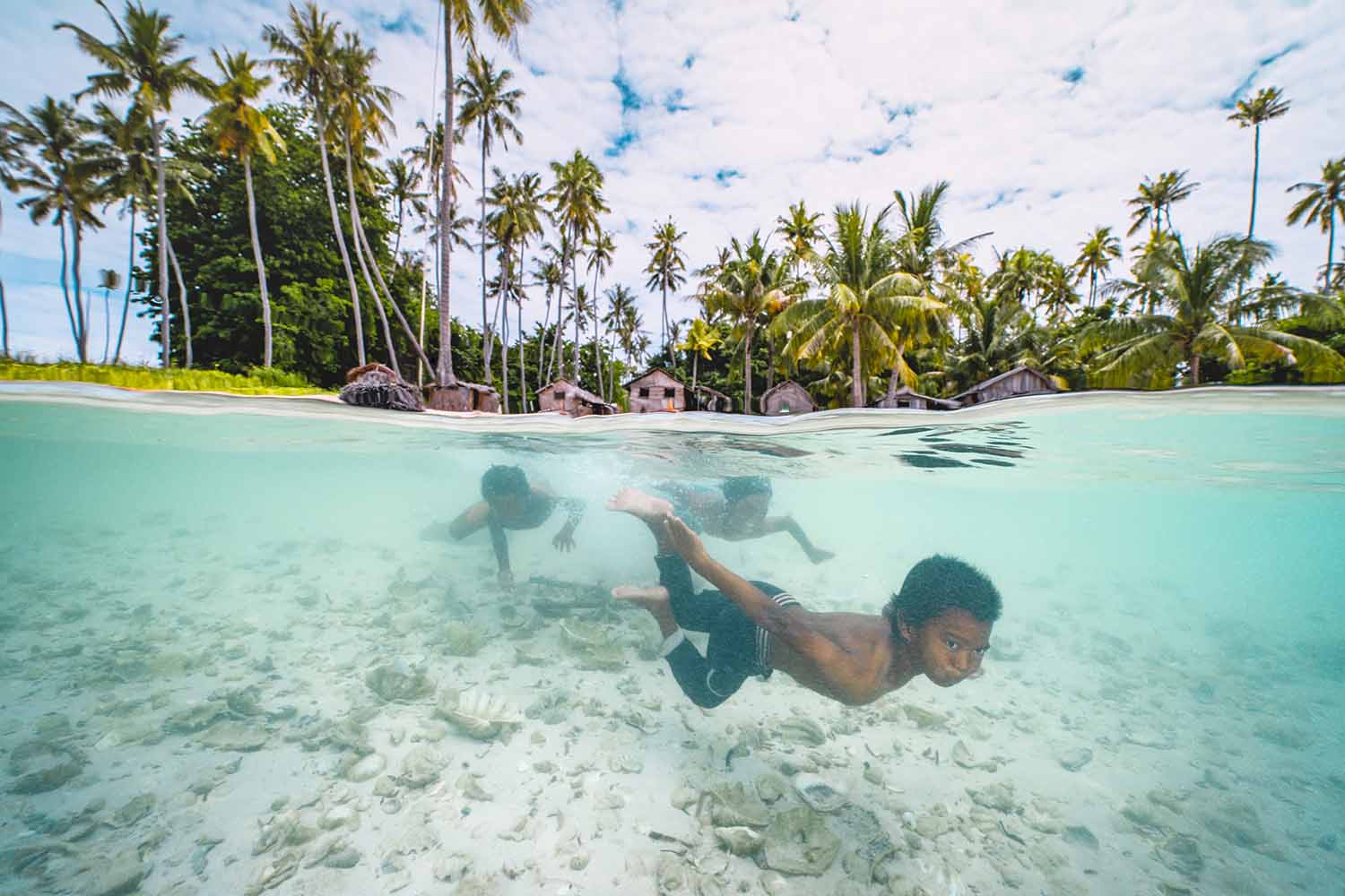 Three children swim underwater. Tropical trees and houses can be seen in the background.