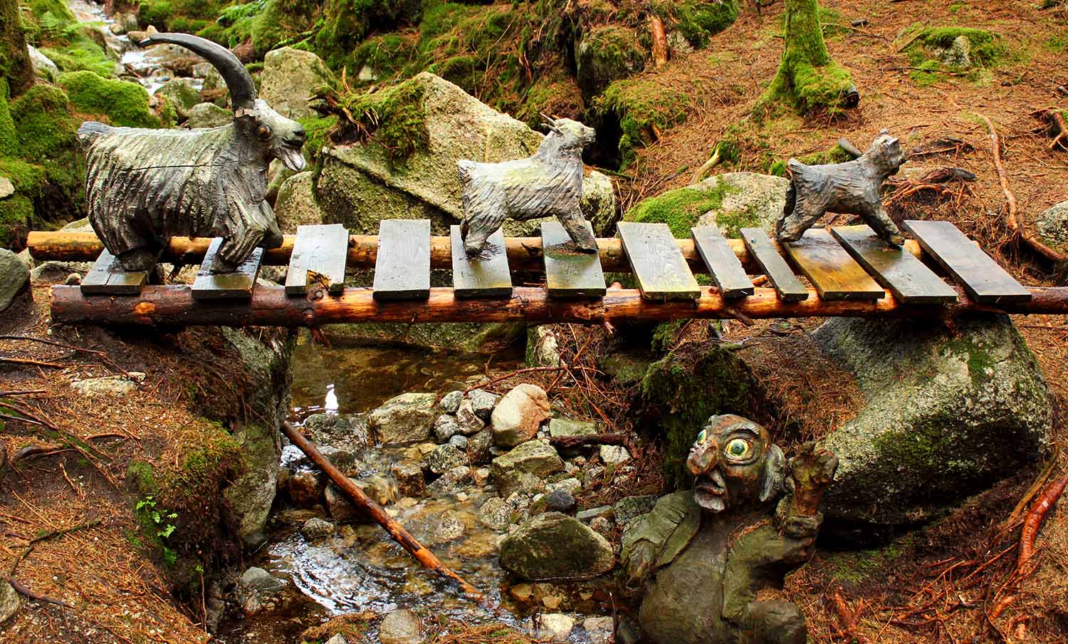 A sculpture of three goats crossing a wooden bridge under which there is a troll.