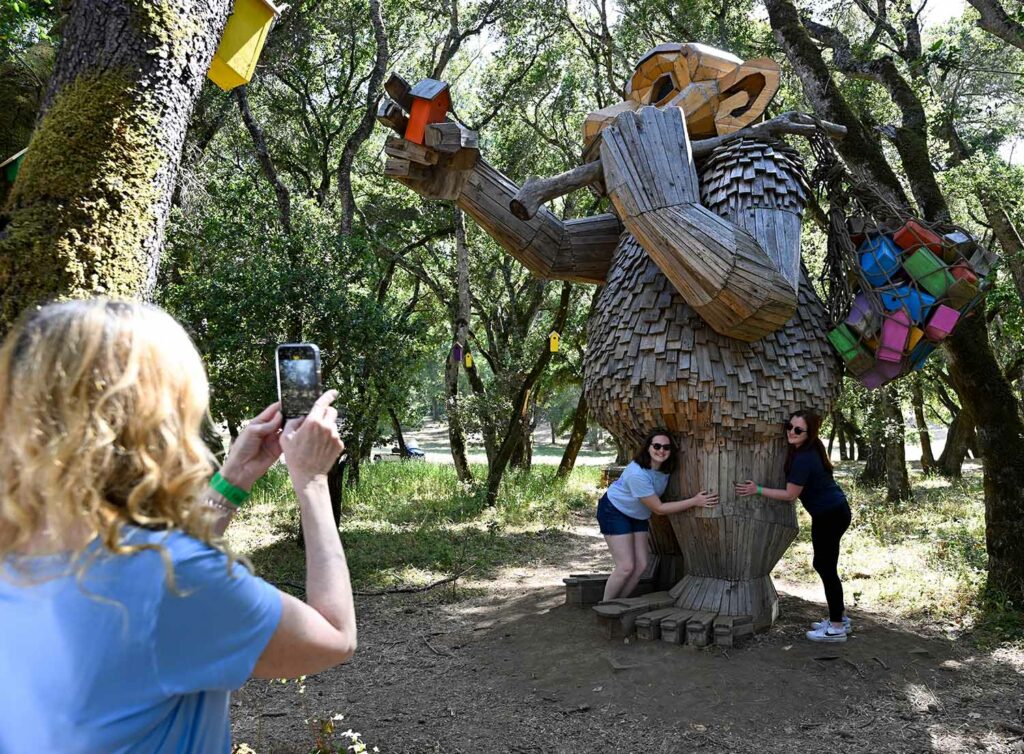 People pose for a photo with a large wooden troll that holds a net full of birdhouses and is about to put a birdhouse on a tree.