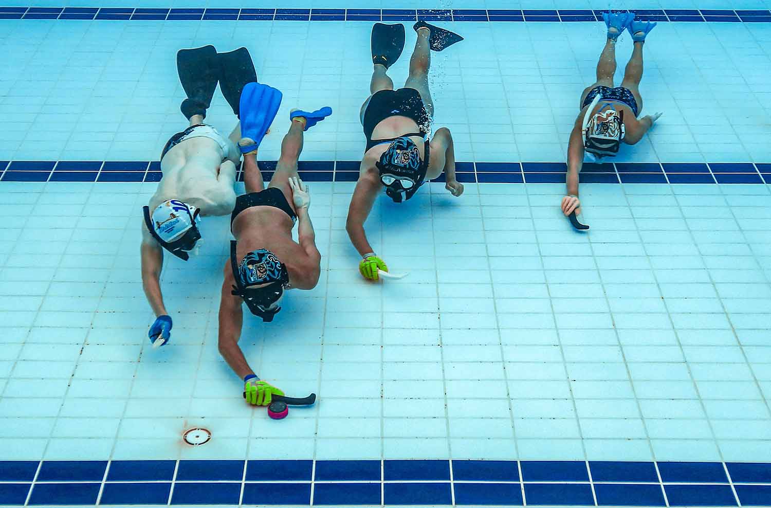 Four swimmers with snorkeling gear hold tiny hockey sticks at the bottom of a pool. One of them is about to push a puck.