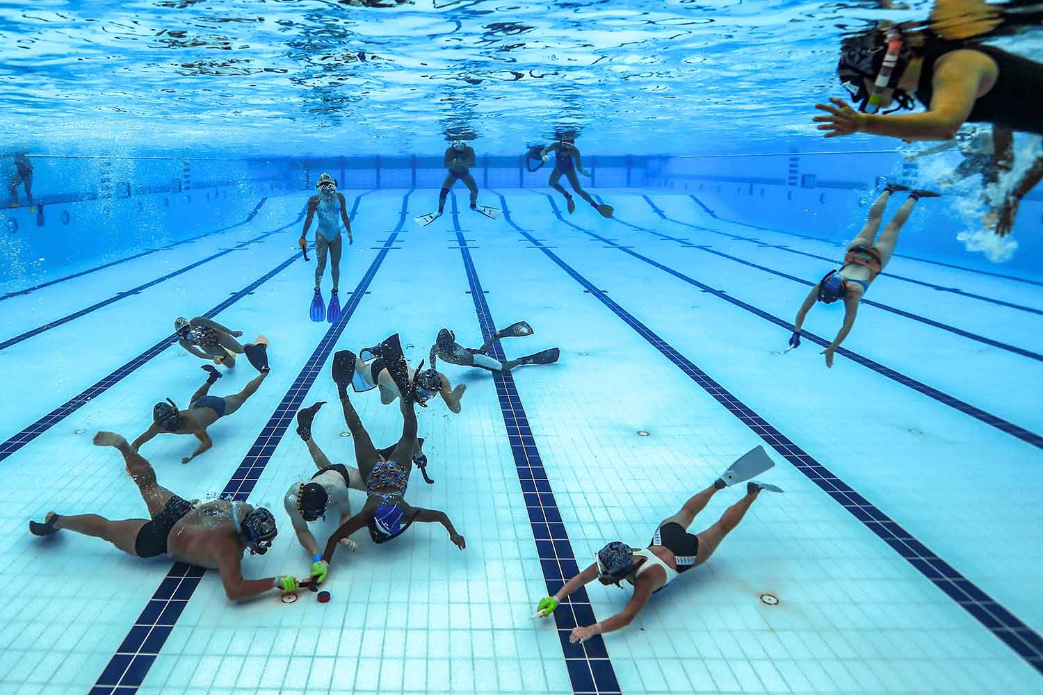 Several people in snorkeling gear and holding tiny hockey sticks gather around or swim toward a puck at the bottom of a pool.