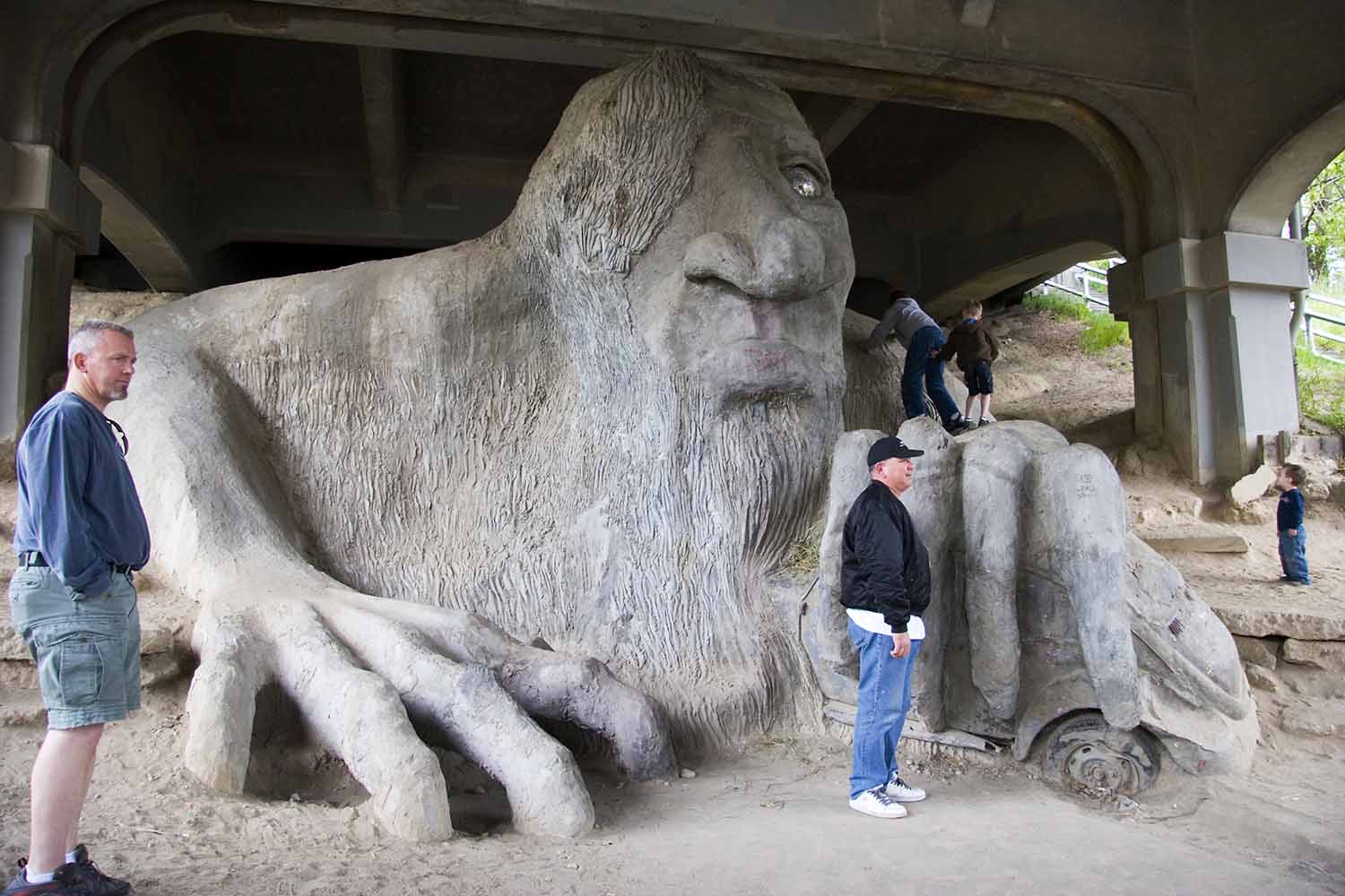 Under a bridge, people pose with a large cement troll that holds a car in one hand.