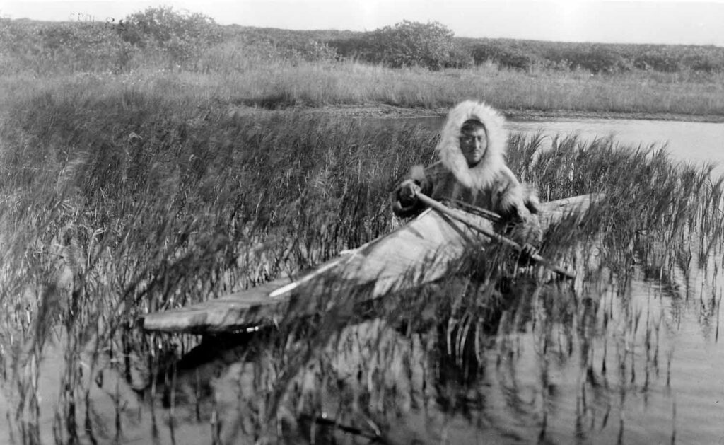A man wearing a coat with a fur lined hood kayaks through a marshy area.