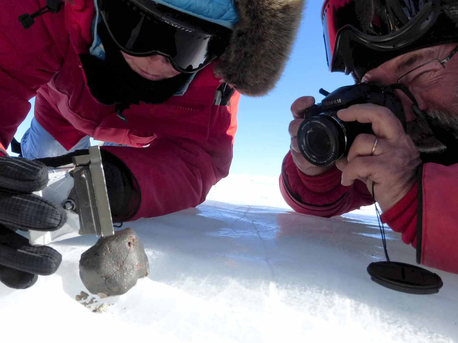 Two people in heavy coats kneel on an icy surface and use a camera and equipment to collect data on a meteorite that is there.