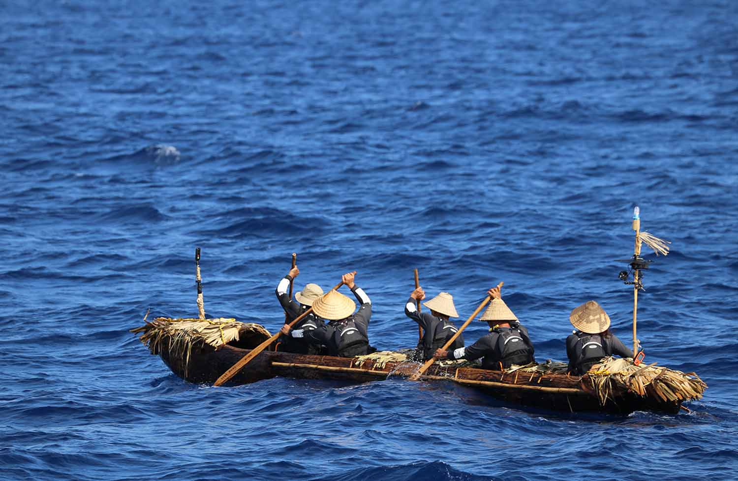 Five people wearing hats and backpacks paddle on a sea in a wooden canoe.