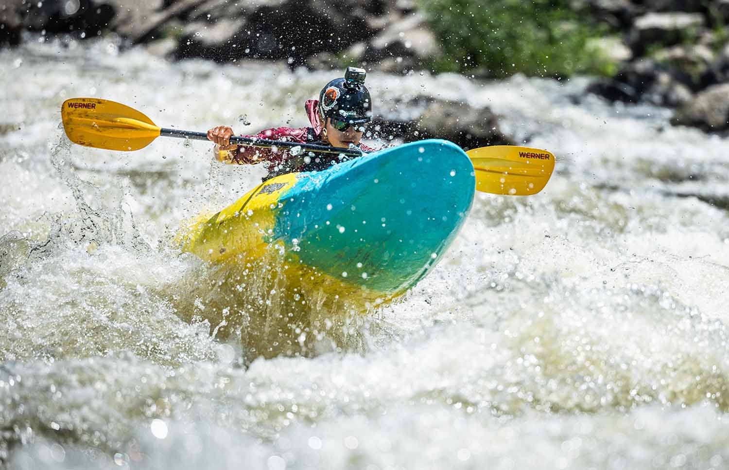 A kayaker with a camera on his helmet uses a paddle to kayak through whitewater.