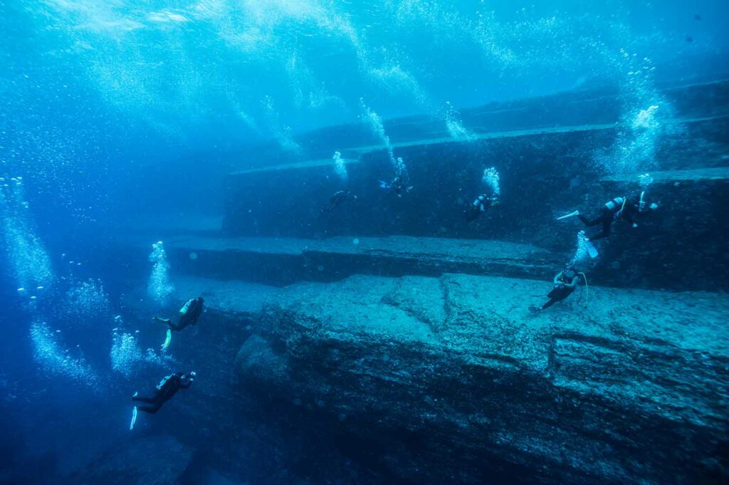 Several scuba divers swim near a huge underwater rock structure that appears to have steps.
