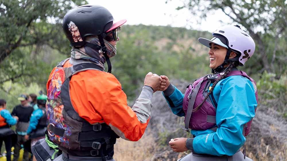 Two kayakers in helmets and other safety gear bump fists with landscape and other kayakers in the background.