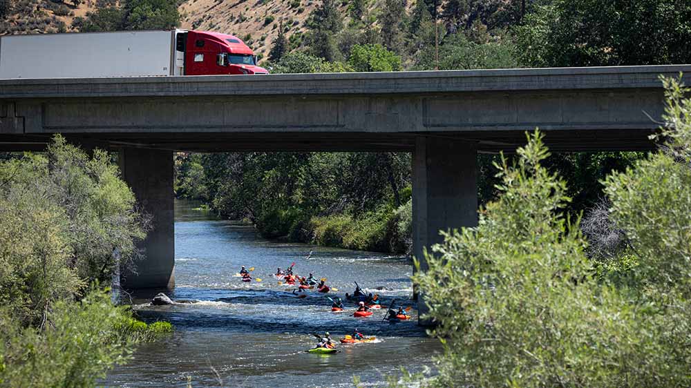 Kayakers are on the river under an overpass on which a truck is traveling.