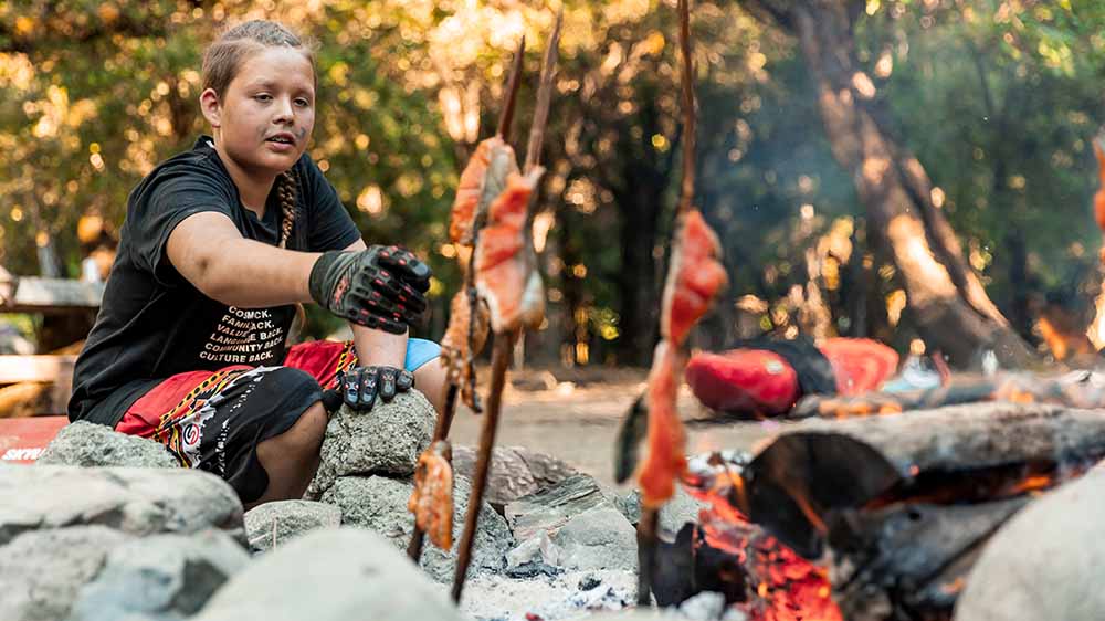 A person is cooking salmon on vertical stakes over an open fire.