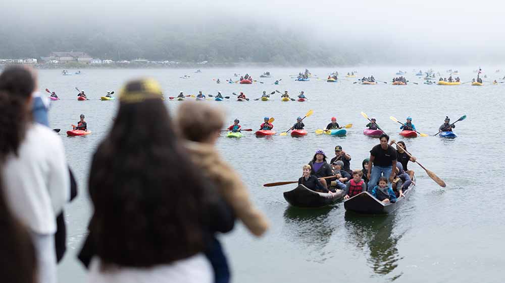 Two people in wooden canoes paddle ahead of many kayakers on the river.