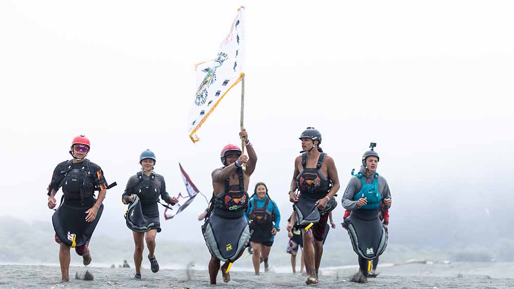 Six people, one carrying a flag, walk on a sandy beach.
