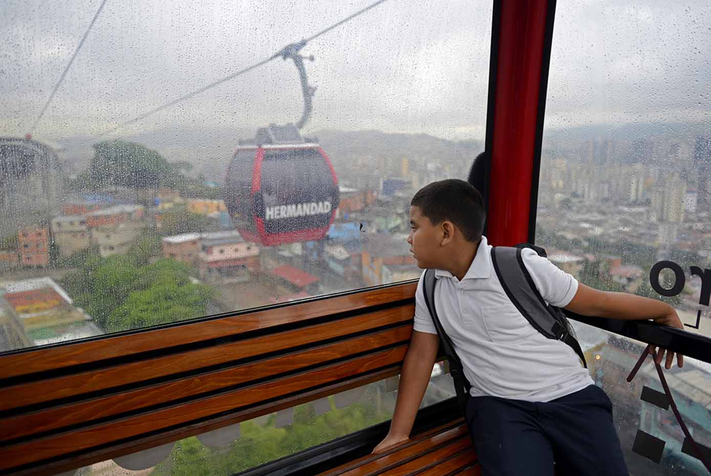 A Venezuelan child with a backpack rides a gondola to school over buildings.