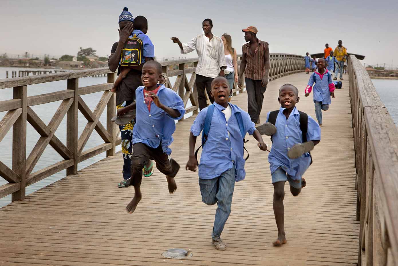 Children wearing school uniforms run across a large wooden bridge while adults walk behind them.