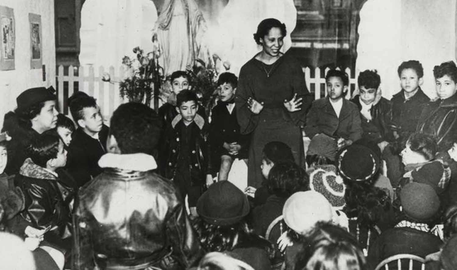 Pura Belpré stands and speaks in a room that is filled with seated children.