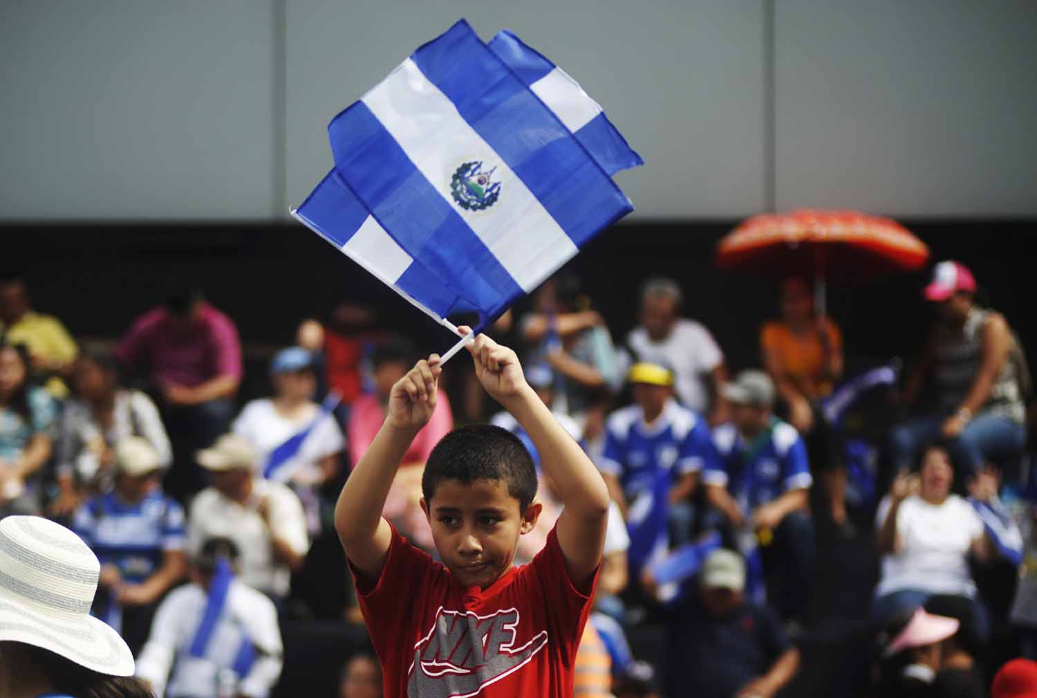 With a crowd of people in the background, a boy waves two flags of El Salvador.