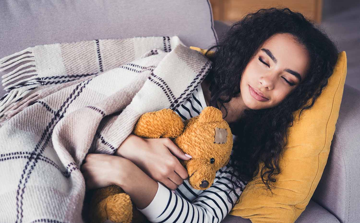 A young woman cuddles a teddy bear as she naps on a couch in a living room.
