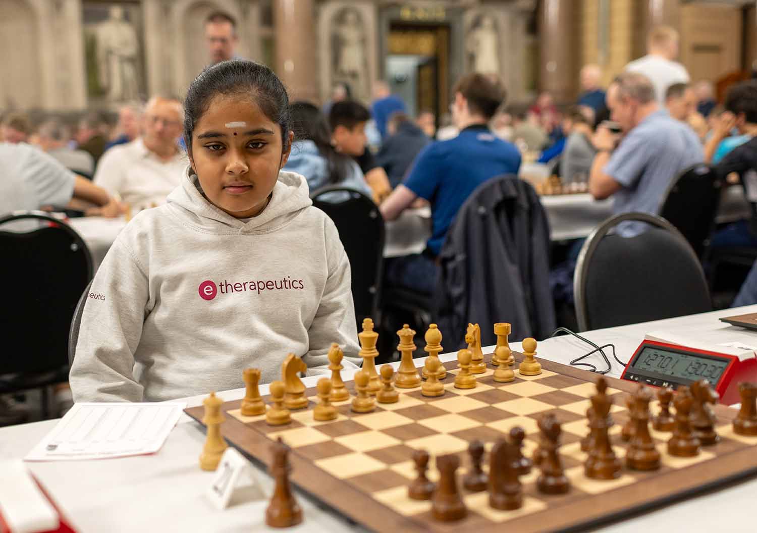 Bodhana Sivananadan sits at a table at a tournament where a chessboard and timer have been set up. She is surrounded by other players.