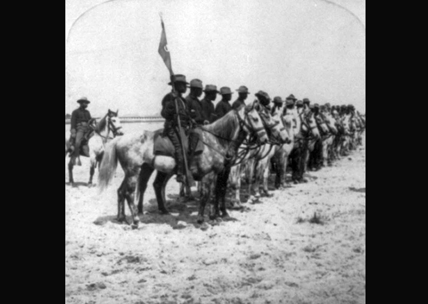 Black soldiers in US military uniforms line up on horseback.