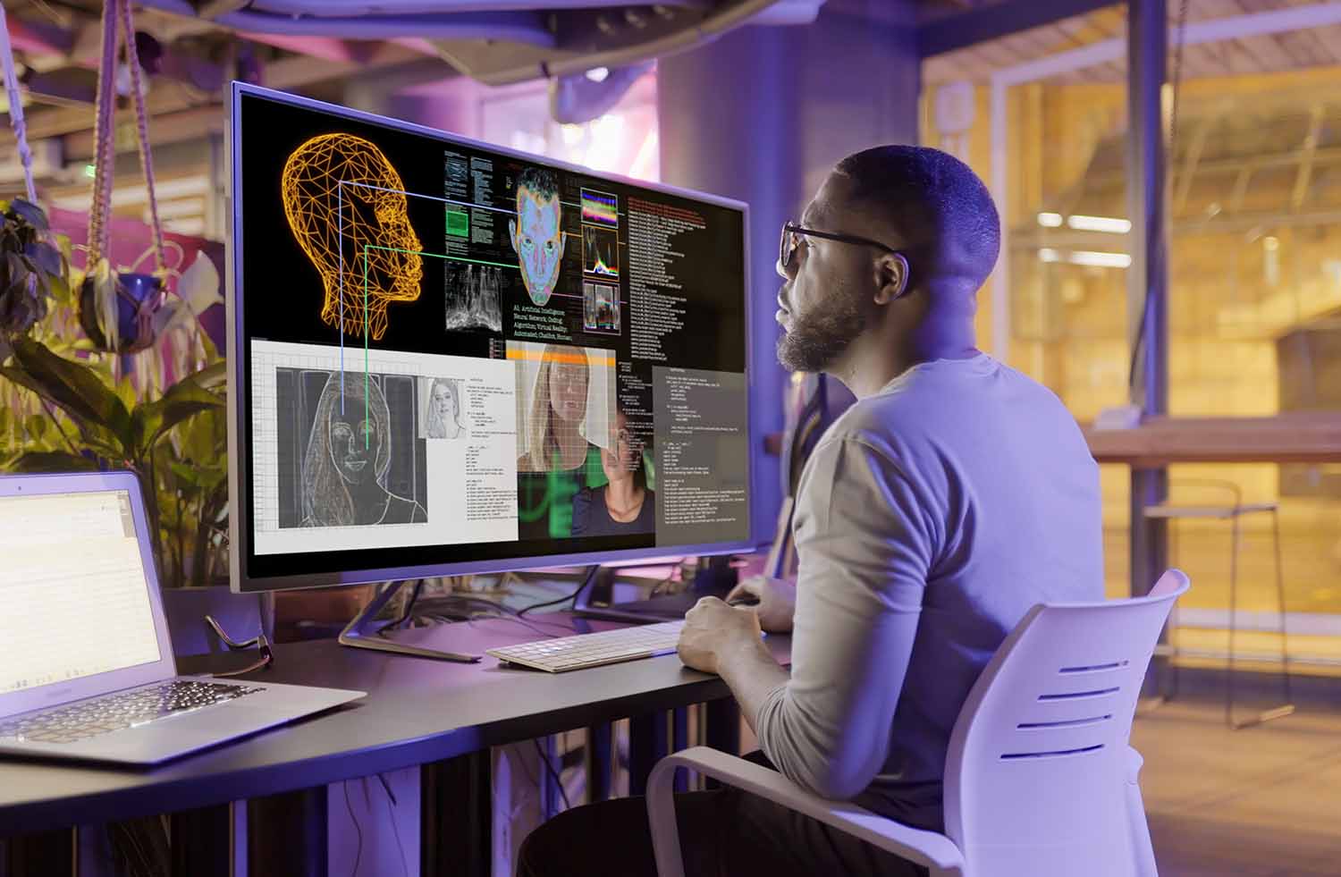 A man sits at a computer and studies many AI generated faces and other images as well as code on a large screen.