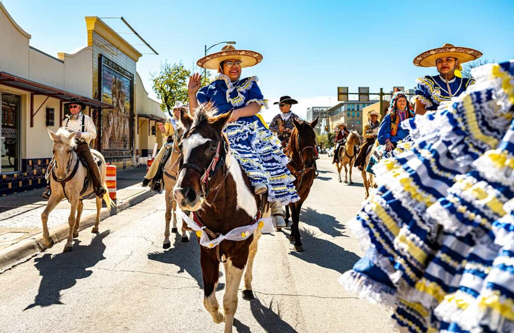 Women and men in colorful dresses and cowboy clothing ride horses down a street.
