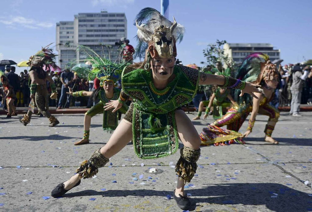 Young people in colorful Indigenous clothing are in dance poses as part of an outdoor parade.