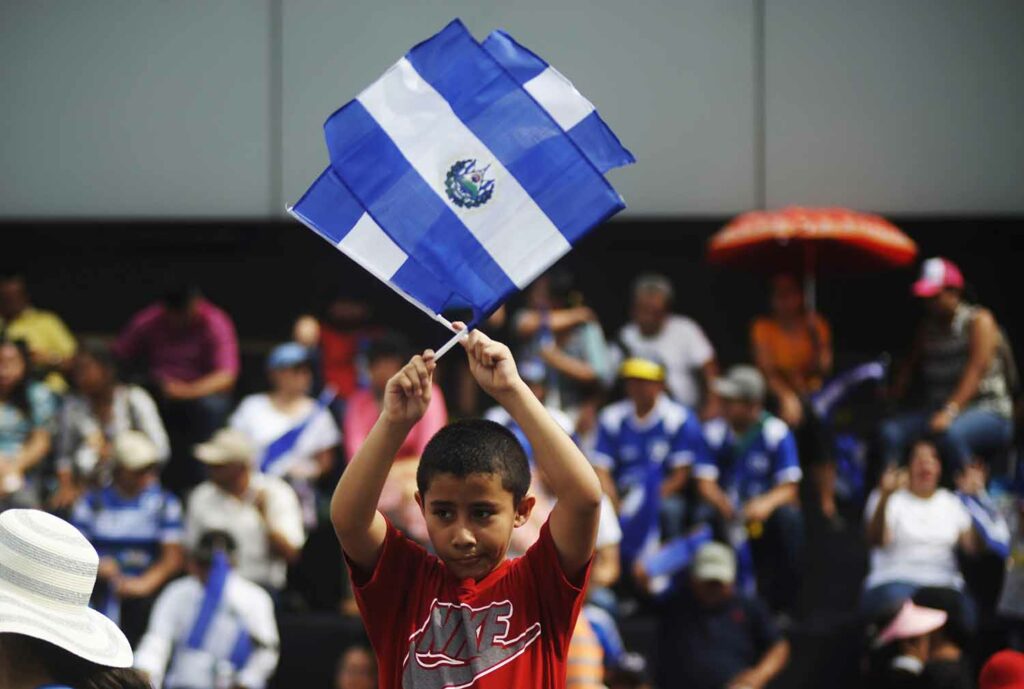 With a crowd of people in the background, a boy waves two flags of El Salvador.