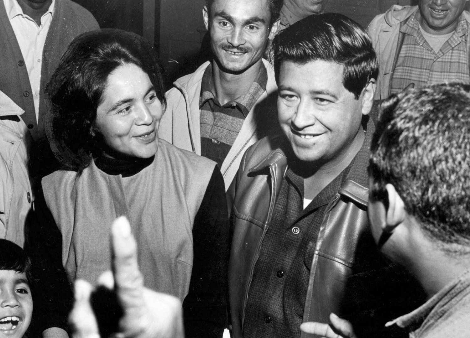 Young Dolores Huerta and Cesar Chavez stand among a group of people in a room.