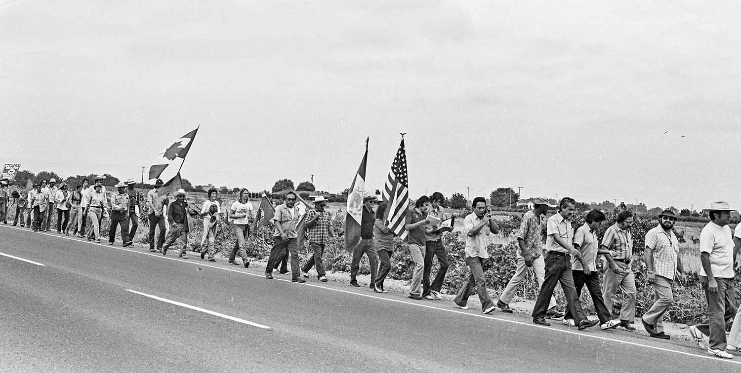 A group of people, some carrying flags and signs, march along the side of a road.