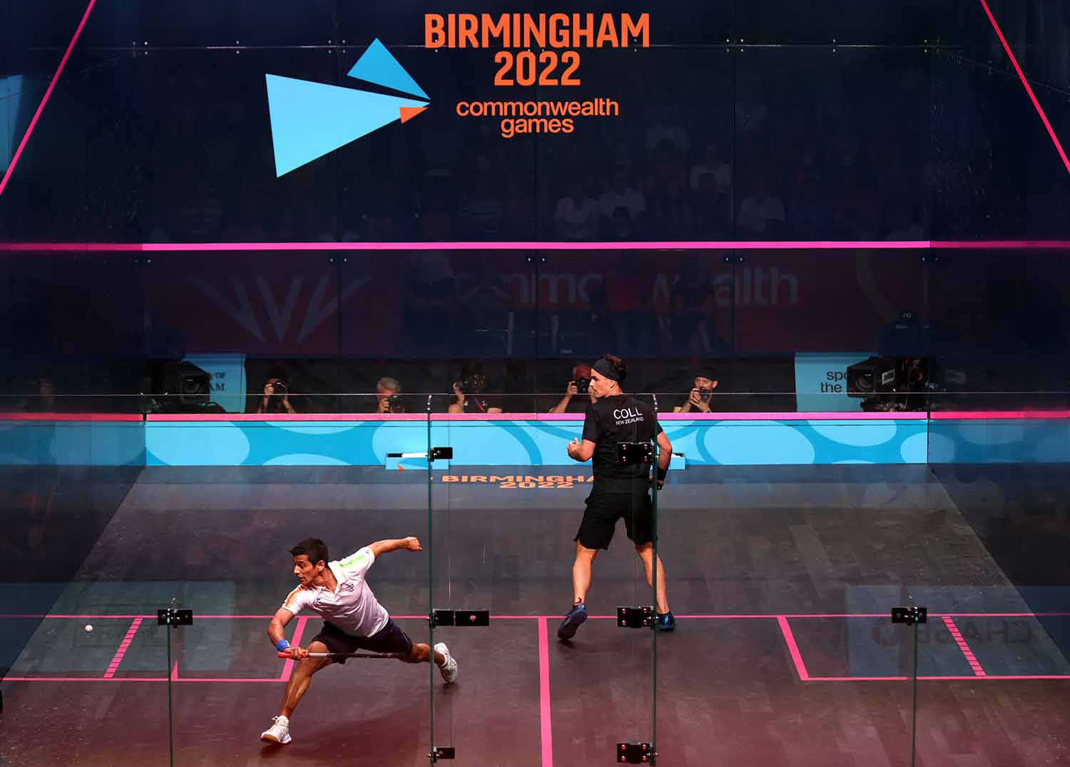 Two players try to hit a ball on a squash court with a sign in the background reading Birmingham 2022 commonwealth games.