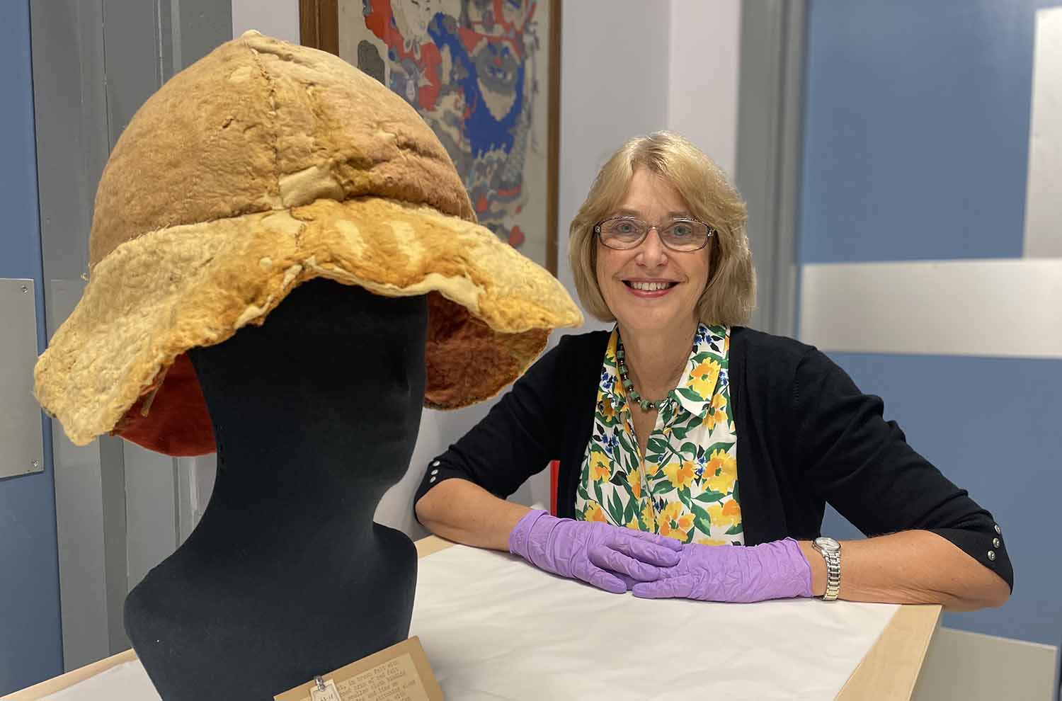 Jacqui Hyman wears purple gloves as she sits next to a felt hat that is on a mannequin.