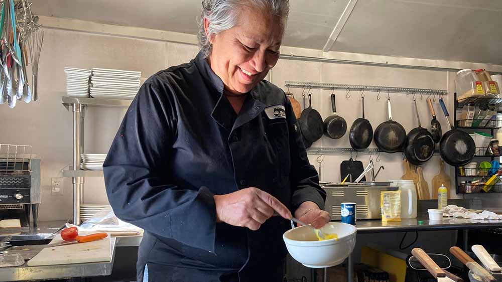 A chef smiles as she stirs food in a bowl as food cooks on a grill.