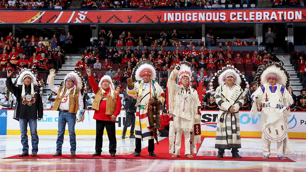 Seven men in headdresses stand on the ice in front of a crowd in an arena in front of the words Indigenous Celebration.