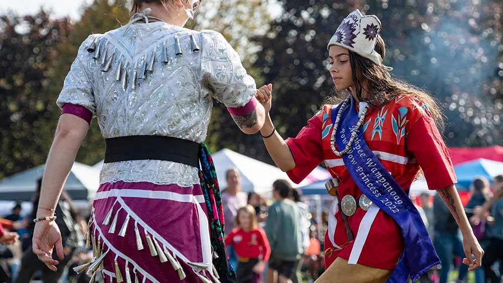 Two people of the Mashpee Wampanoag in traditional dress dance at an outdoor event.